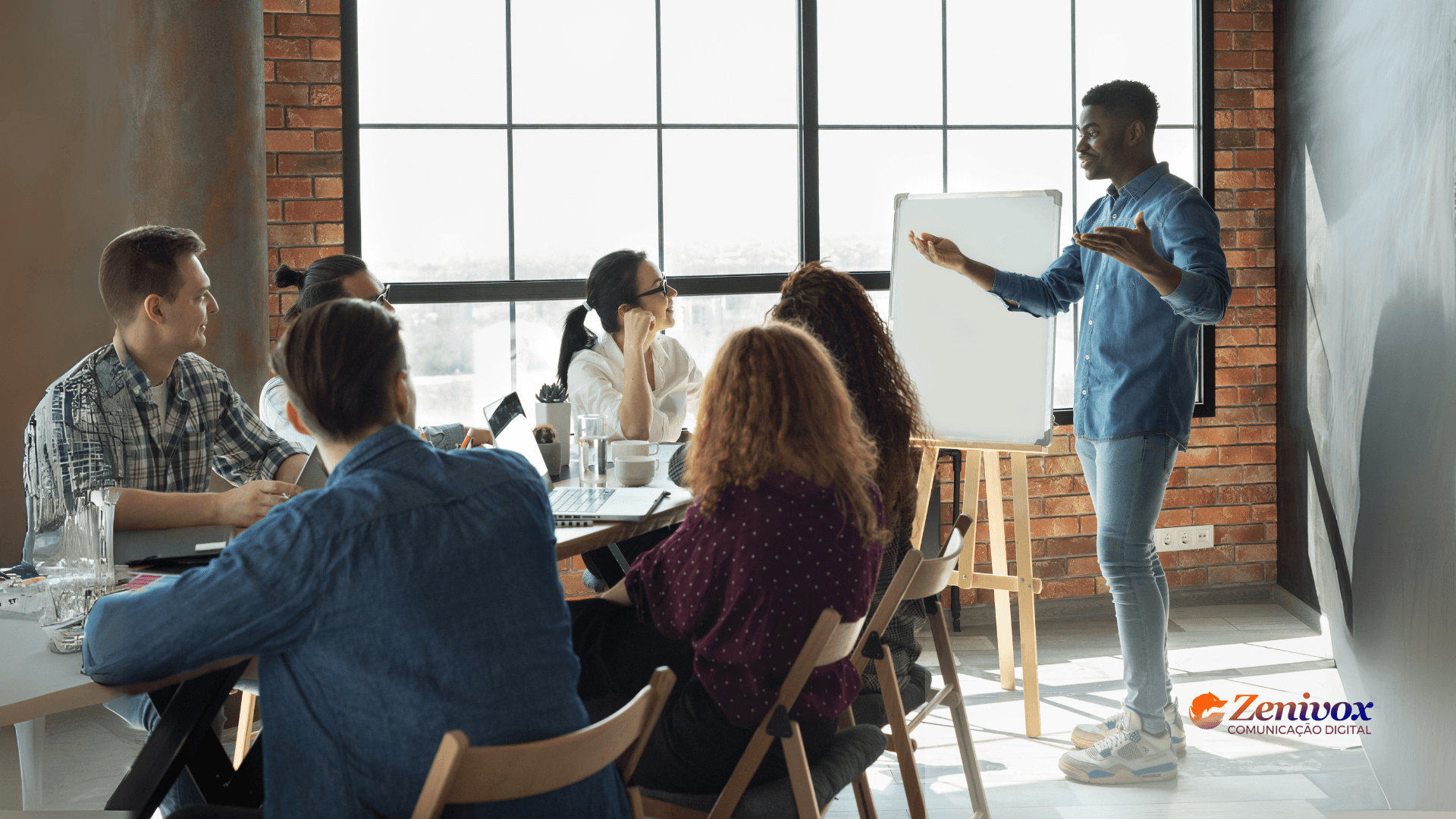 Palestrante em pé conduzindo uma apresentação para uma equipe reunida ao redor de uma mesa em ambiente moderno, demonstrando técnicas de liderança carismática para inspirar e engajar o grupo.