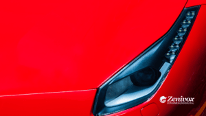 Imagem de um close no farol dianteiro de um carro esportivo vermelho, destacando linhas elegantes e modernas. O fundo é predominantemente vermelho brilhante, transmitindo luxo e performance. Simbolizando o marketing de segmentação da Ferrari. No canto inferior direito, o logotipo da Zenivox Comunicação Digital é visível.