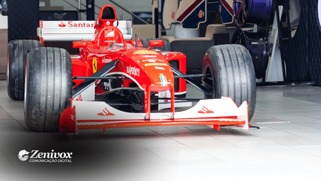 Imagem de um carro de Fórmula 1 da Ferrari, com destaque para seu design aerodinâmico em vermelho vibrante e os logotipos de marcas parceiras no chassi. O ambiente sugere um showroom ou espaço promocional, reforçando a conexão entre a Ferrari e marcas de luxo. No canto inferior esquerdo, está presente o logotipo da Zenivox Comunicação Digital.