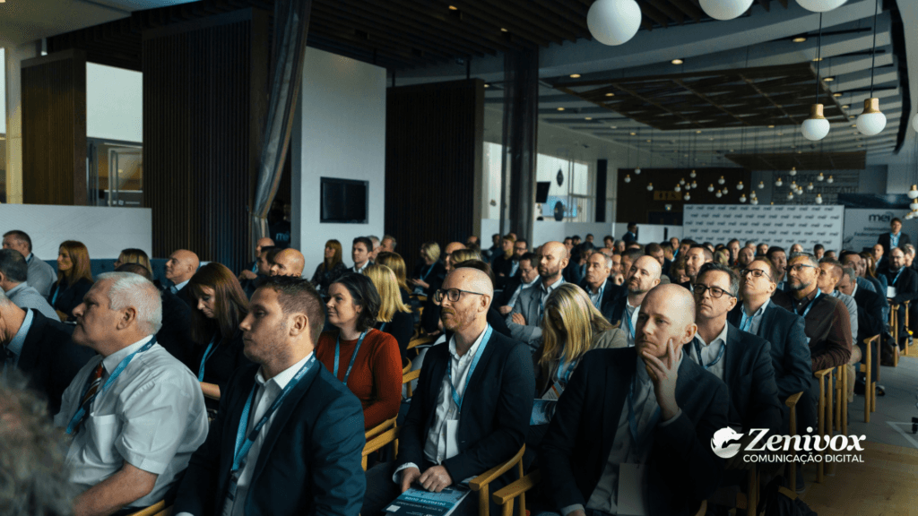 Uma sala cheia de profissionais observa atentamente a uma apresentação em um evento corporativo. As pessoas estão sentadas, usando roupas formais e usando crachás, simbolizando um ambiente de alinhamento estratégico e colaboração. A logo da Zenivox Comunicação Digital aparece no canto inferior direito, destacando seu papel em estratégias de marketing integradas.