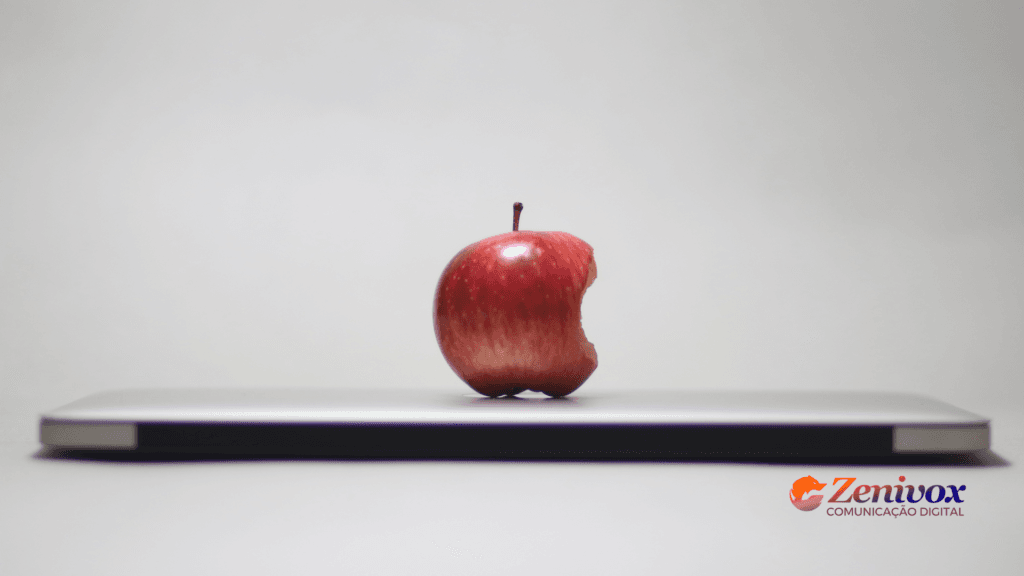Uma maçã vermelha mordida sobre um laptop prateado fechado, remetendo à identidade visual da Apple. A imagem representa o conceito de posicionamento de marca e a rivalidade entre Apple e Samsung no mercado de tecnologia. No canto inferior direito, o logotipo da Zenivox Comunicação Digital reforça a identidade visual.