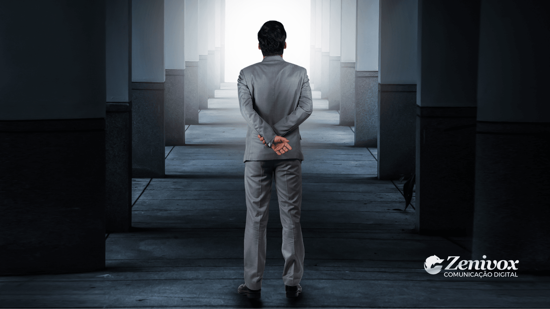 Homem de terno cinza visto de costas, caminhando por um corredor simétrico com iluminação ao fundo, representando a jornada estratégica e desafios do Gerente de Vendas. A imagem transmite profissionalismo, tomada de decisão e busca por resultados extraordinários.
