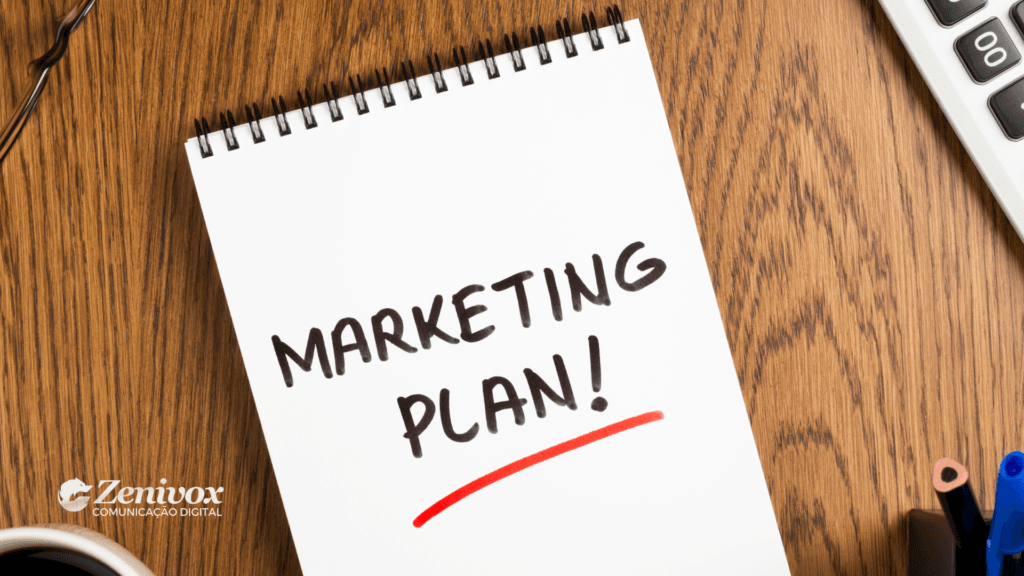 Bloco de anotações com a frase "Marketing Plan!" escrita em letras grandes e sublinhada em vermelho. O caderno está sobre uma mesa de madeira, acompanhado de uma caneca de café, uma calculadora e materiais de escritório. No canto inferior esquerdo, aparece o logotipo da Zenivox Comunicação Digital.