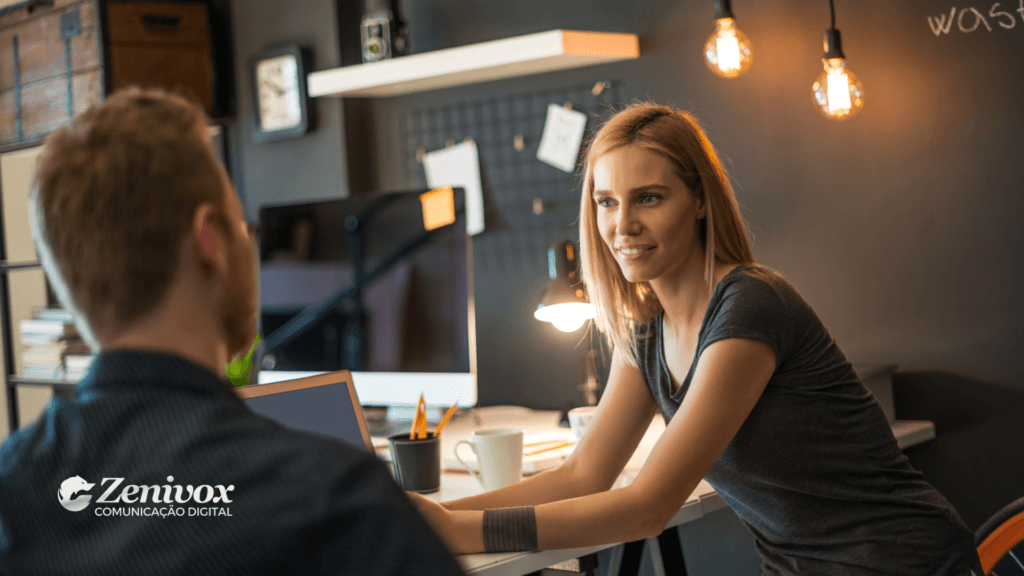 Cena em ambiente de escritório criativo, com uma mulher e um homem em uma reunião colaborativa. A mulher está atenta e engajada na conversa, representando a importância do diálogo entre líderes e equipes para desenvolver uma visão de negócios eficaz. Ao fundo, as luminárias pendentes criam uma atmosfera moderna e acolhedora. O logotipo da Zenivox Comunicação Digital está localizado no canto inferior esquerdo.