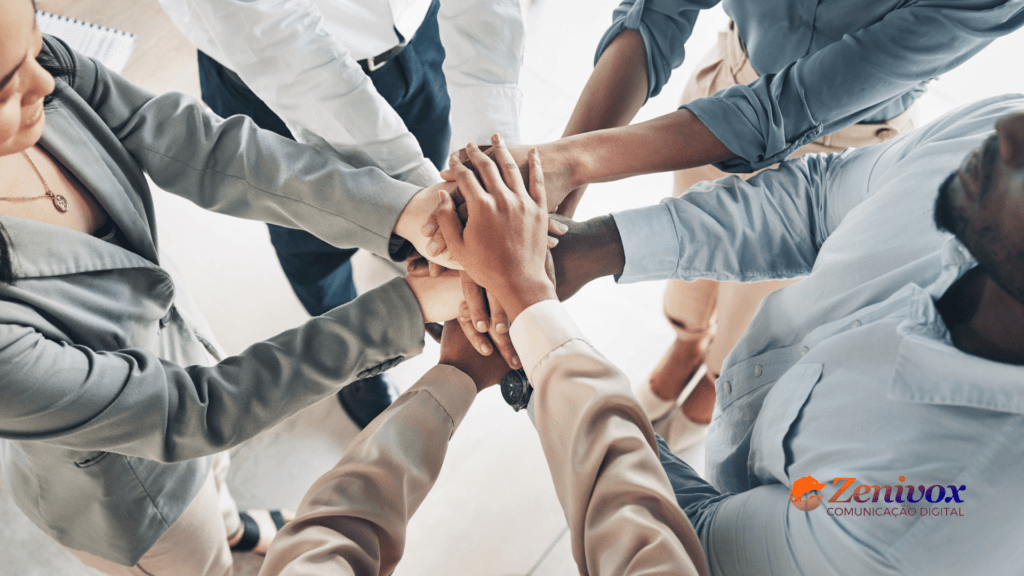 Equipe de colaboradores em um gesto de união, com todas as mãos sobrepostas no centro, simbolizando o alinhamento e o comprometimento com os objetivos empresariais. A imagem representa a importância da comunicação eficaz para garantir que todos compreendam e se conectem com a visão de negócios. O logotipo da Zenivox Comunicação Digital está localizado no canto inferior direito.