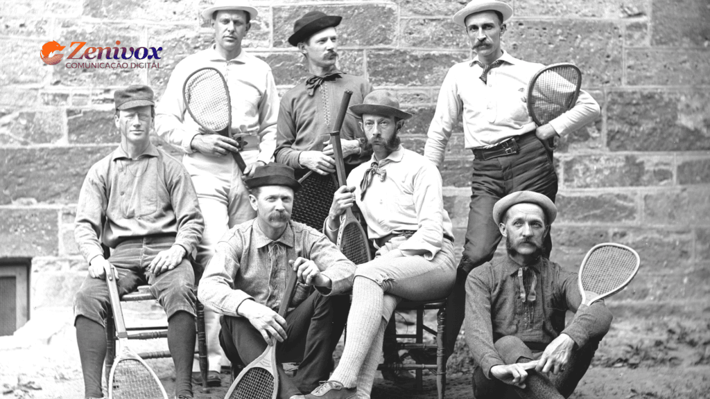Imagem histórica em preto e branco de sete homens vestidos com trajes antigos, típicos do século XIX, posando com raquetes de tênis em frente a uma parede de pedra. Cada homem exibe expressões sérias e posturas distintas, com alguns sentados e outros em pé. A imagem remete ao contexto esportivo clássico, destacando a origem e a tradição desse universo.