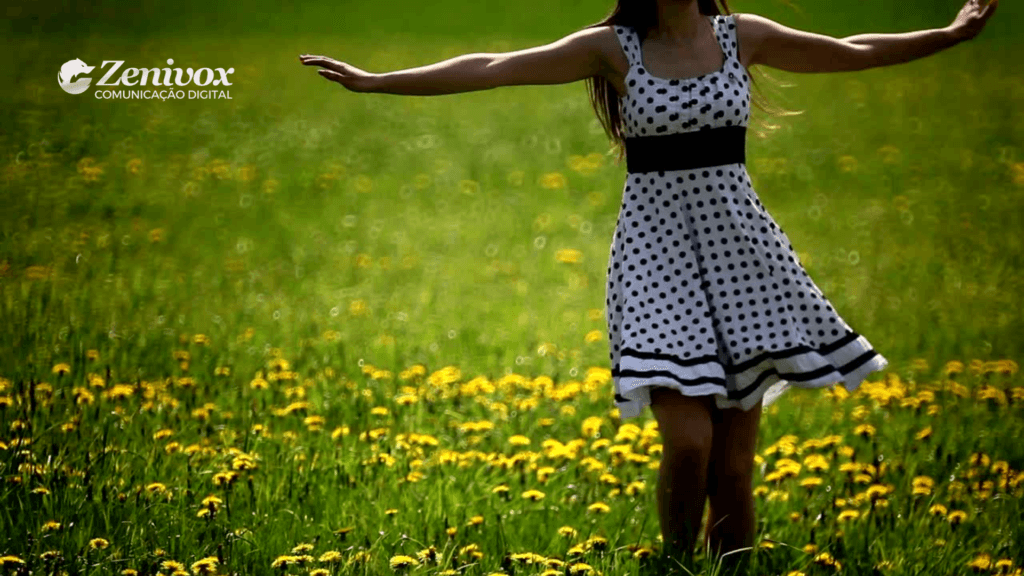 Imagem de uma mulher vestida com um vestido branco de bolinhas pretas, dançando livremente em um campo repleto de flores amarelas. O logotipo da Zenivox Comunicação Digital está presente no canto superior esquerdo. A imagem simboliza liberdade, conexão com a natureza e bem-estar, remetendo ao conceito de sustentabilidade e práticas ecológicas.