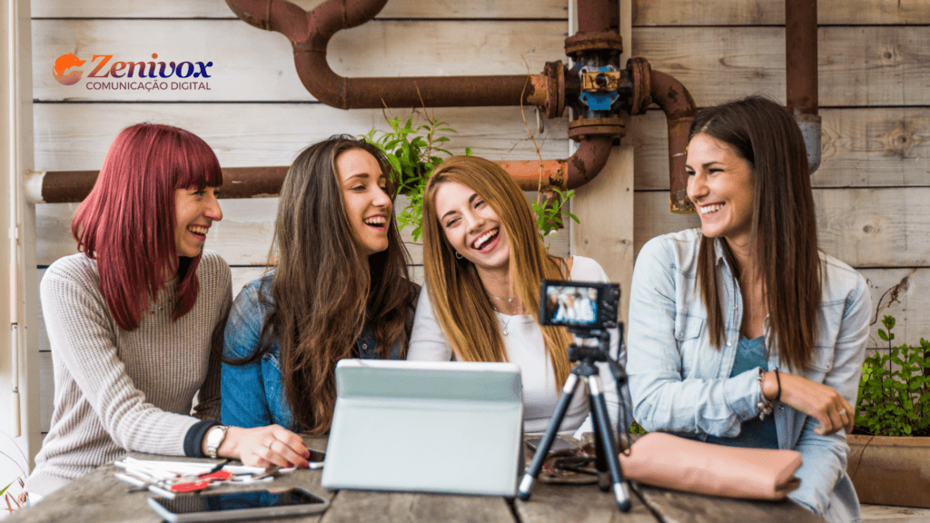 Quatro jovens mulheres sentadas em uma mesa ao ar livre, rindo e se divertindo durante uma gravação para uma câmera posicionada sobre um tripé. Na mesa, há um tablet, cadernos e celulares, informando um momento de criação de conteúdo colaborativo e espontâneo para o marketing de influência. O logotipo da Zenivox está presente no canto superior esquerdo.