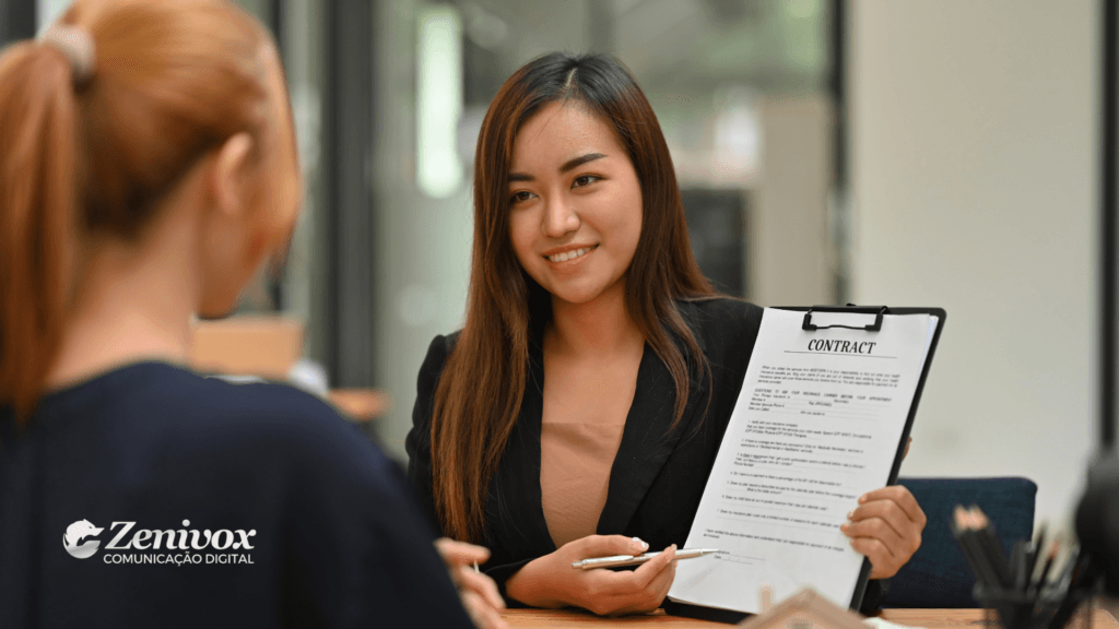 Uma mulher sorridente, vestida com um blazer preto, apresenta um contrato a uma cliente enquanto segura uma caneta, sugerindo um fechamento de negociação. A imagem transmite profissionalismo e a importância de dominar técnicas de negociação para alcançar resultados positivos. O logo da Zenivox Comunicação Digital está visível no canto inferior esquerdo.