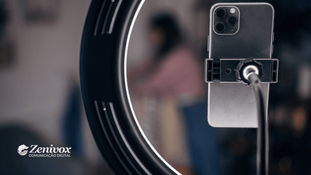 ma imagem focada em um smartphone montado em um suporte para gravação de vídeo, cercado por um ring light aceso. No fundo desfocado, uma pessoa é visível, revelando um ambiente de criação de conteúdo digital. O logotipo da Zenivox está posicionado no canto inferior esquerdo, baixando uma abordagem profissional para marketing digital.
