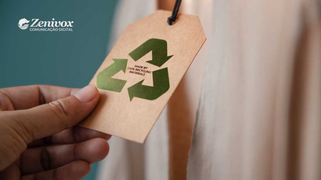 Imagem de uma etiqueta de papel reciclado com o símbolo de reciclagem em destaque e a mensagem "MADE BY 100% RECYCLING MATERIALS" (Feito com 100% de materiais reciclados). A etiqueta está sendo segurada por uma mão e está presa a uma peça de roupa clara ao fundo. O logotipo da Zenivox Comunicação Digital está posicionado no canto superior esquerdo, destacando a conexão com práticas ecológicas e sustentáveis.