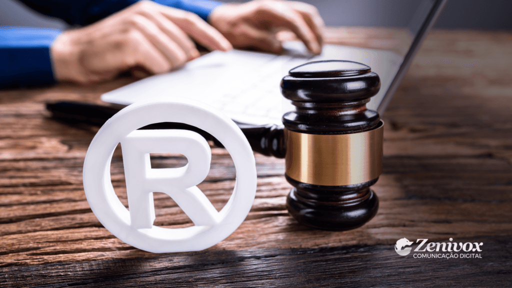 Imagem representando conceitos de branding pessoal e propriedade intelectual. Em primeiro plano, há um símbolo de marca registrada (®) e um martelo de juiz, ambos sobre uma mesa de madeira, simbolizando a proteção legal e a construção de uma marca sólida. Ao fundo, uma pessoa usando camisa azul está digitando em um notebook, remetendo à parte estratégica e digital da construção da imagem profissional. No canto inferior direito, o logotipo da Zenivox Comunicação Digital está visível.
