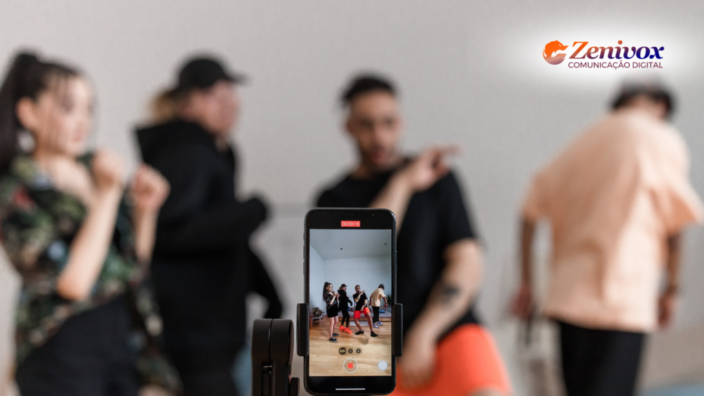 Smartphone grava um grupo de jovens dançando juntos em um ambiente interno. A câmera foca na tela do celular, que exibe a gravação, enquanto os dançarinos ao fundo estão desfocados. No canto superior direito, aparece o logo da Zenivox Comunicação Digital.