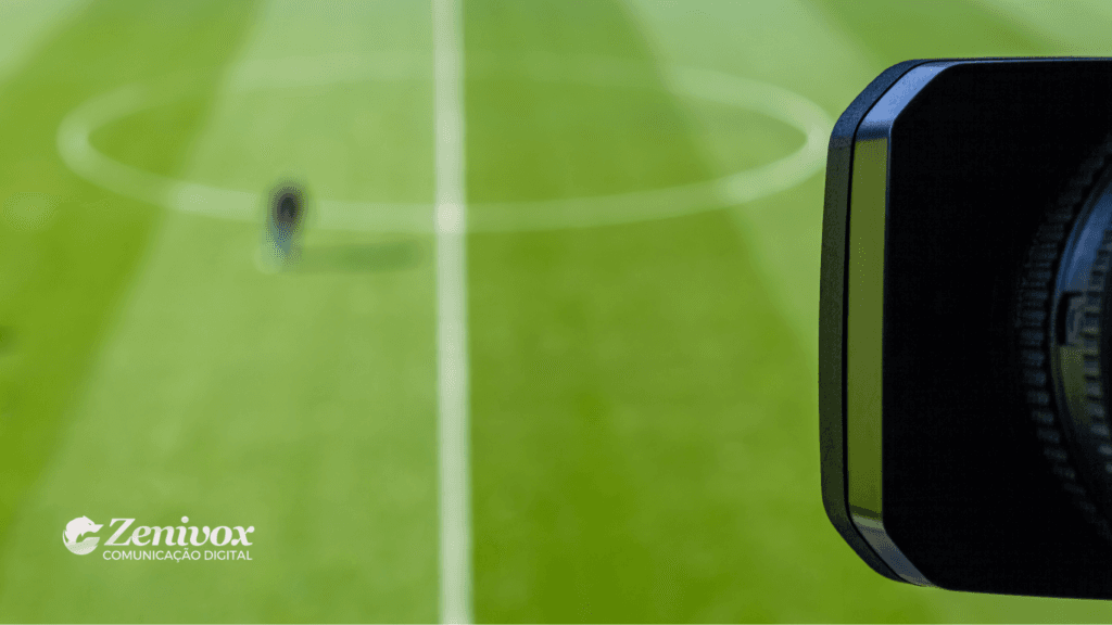 Imagem desfocada de um campo de futebol com uma câmera de vídeo em primeiro plano à direita. No centro do campo, um indivíduo aparece embaçado. No canto inferior esquerdo está o logotipo da Zenivox Comunicação Digital.