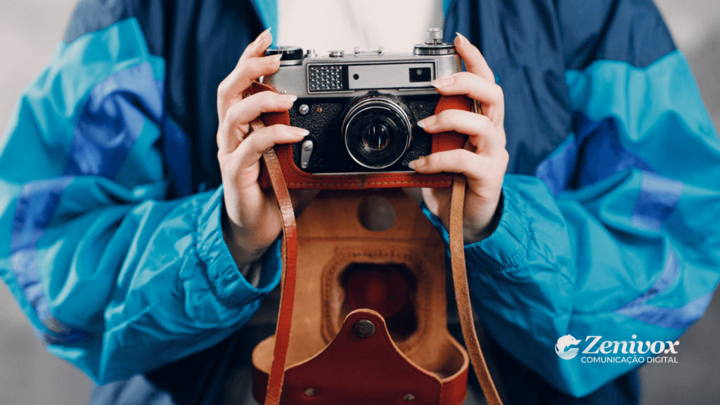 Pessoa com jaqueta azul estilo retrô segurando uma câmera analógica vintage com case de couro aberta, remetendo à fotografia tradicional e ao resgate de memórias afetivas.