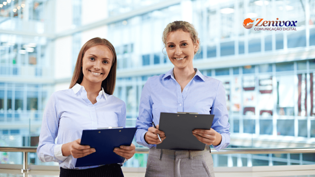 Duas mulheres sorridentes usam roupas sociais e seguram pranchetas em um ambiente corporativo moderno, com fundo envidraçado. Elas transmitem confiança e profissionalismo. No canto superior direito da imagem, aparece o logotipo da ZeniVox Comunicação Digital.