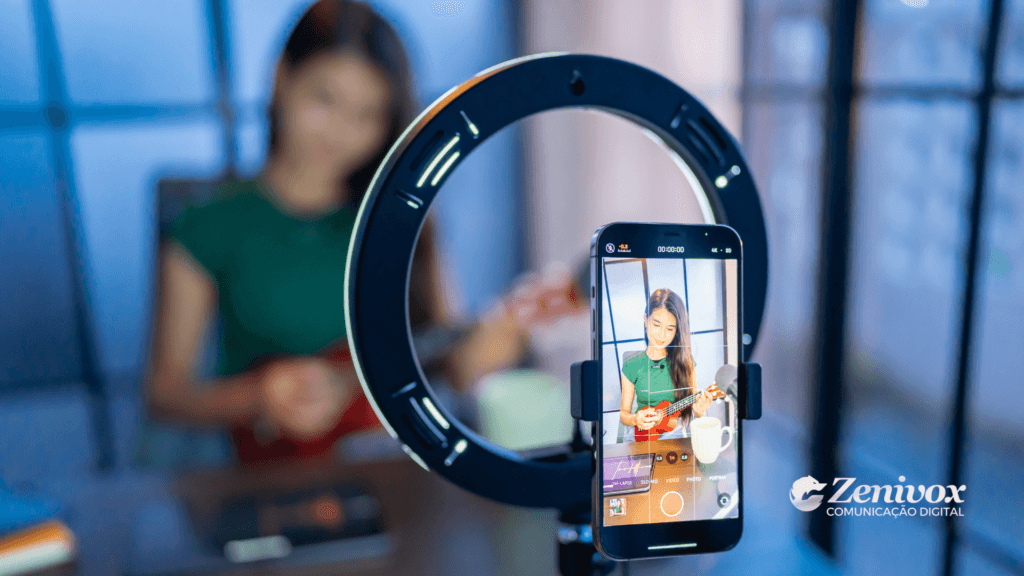Jovem grava um vídeo tocando um ukulele, posicionada em frente a um ring light com um smartphone registrando a cena. A tela do celular mostra a imagem focada nela, enquanto o fundo permanece desfocado. No canto inferior direito, está o logo da Zenivox Comunicação Digital.
