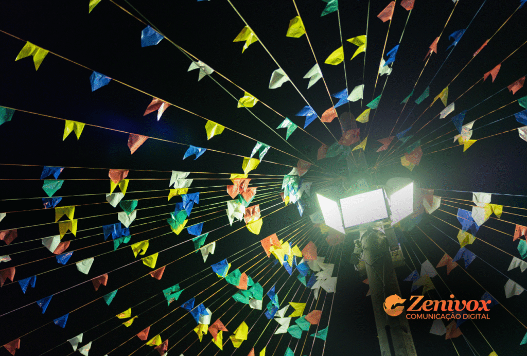 Imagem noturna com bandeirolas coloridas de festa junina amarradas em formato radial, partindo de um poste de luz central. A atmosfera celebra o São João com intensidade simbólica, cultural e econômica atraves do market share. No canto inferior direito, a marca Zenivox Comunicação Digital está visível.
