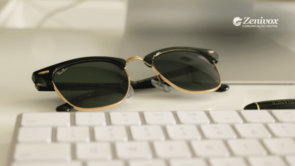 Óculos de sol da marca Ray-Ban em destaque sobre uma mesa branca. O design clássico com armação preta e dourada está em foco ao lado de um teclado branco e uma caneta preta com detalhes dourados. Ao fundo, o logotipo da Zenivox Comunicação Digital aparece no canto superior direito da imagem.