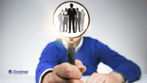 Pessoa segura uma lupa voltada para a câmera, com a lente ampliando a imagem de silhuetas de profissionais de negócios. A imagem simboliza a identificação e valorização de talentos para uma gestão por competência. No canto inferior esquerdo, está o logotipo da Zenivox Comunicação Digital.