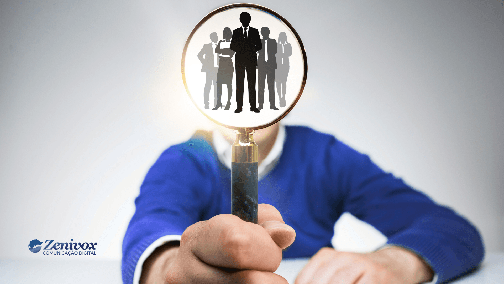 Pessoa segura uma lupa voltada para a câmera, com a lente ampliando a imagem de silhuetas de profissionais de negócios. A imagem simboliza a identificação e valorização de talentos para uma gestão por competência. No canto inferior esquerdo, está o logotipo da Zenivox Comunicação Digital.