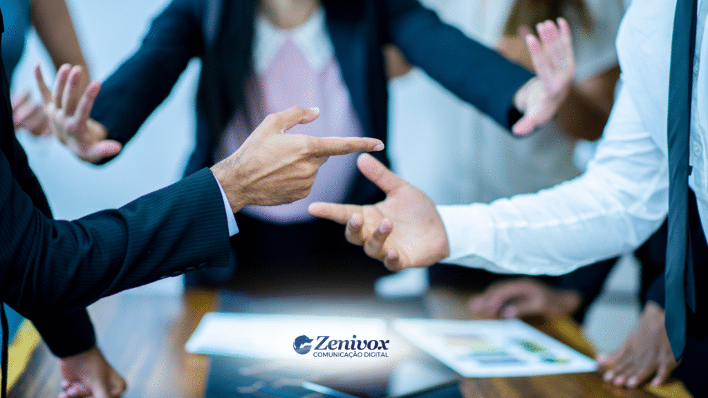 Cena de uma discussão acalorada em ambiente corporativo. Diversas mãos em gestos de apontar e interromper indicam um conflito entre colegas de trabalho. A imagem transmite tensão e urgência e gestão de conflitos, com destaque para o embate verbal e corporal. Na parte inferior, o logotipo da Zenivox Comunicação Digital.