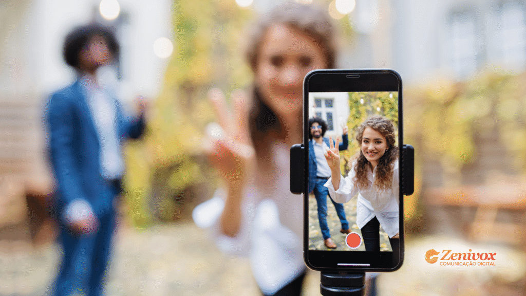 Mulher sorridente fazendo gesto de paz enquanto grava um vídeo com smartphone preso a um tripé. O foco da imagem está na tela do celular, que mostra com nitidez a gravação, enquanto o fundo está desfocado, representando ferramentas de ia para criar . O logo da Zenivox Comunicação Digital aparece no canto inferior direito.