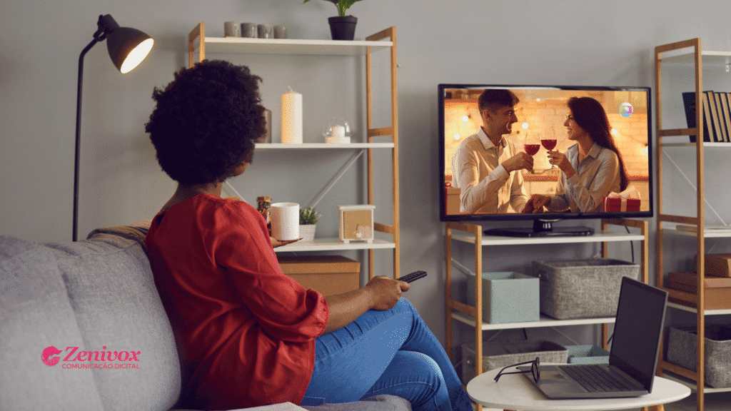 Mulher sentada em um sofá assistindo à televisão em uma sala aconchegante. Na TV, um casal brinda com taças de vinho em uma cena romântica de novela exibida na Globo, com um presente visível ao lado — referência sutil à presença de marca.
