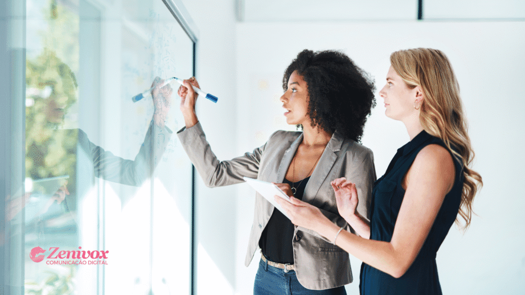 Duas mulheres em ambiente corporativo, uma escrevendo em um quadro de vidro com marcador azul e a outra segurando um tablet, ambas discutindo estratégias.